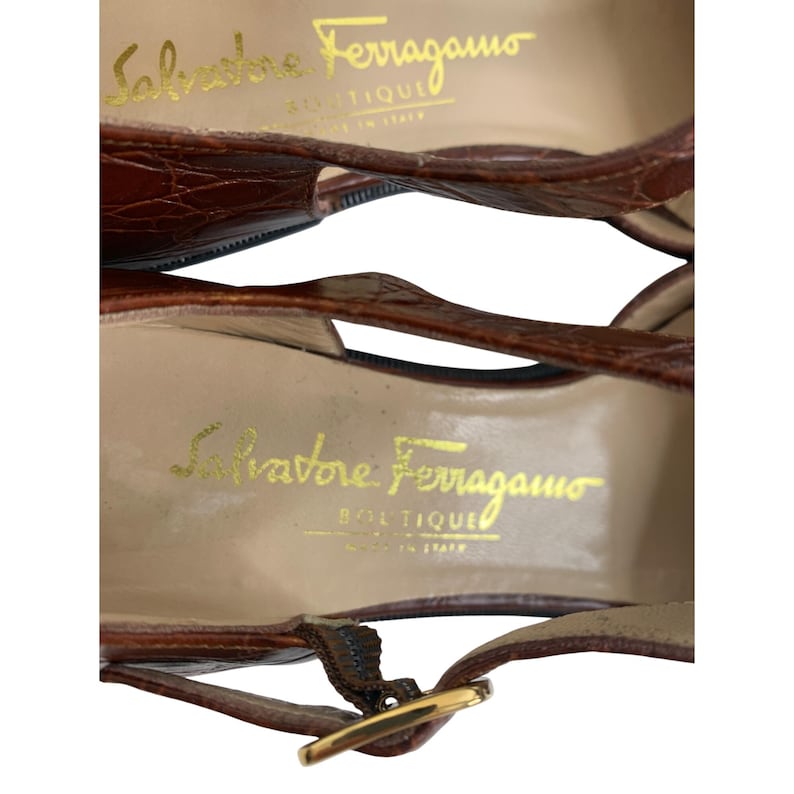 May include: Close-up of two brown leather Salvatore Ferragamo Boutique sandals. The sandals have a textured, crocodile-like pattern. The brand name is printed in gold on the insole. The sandals have a gold buckle.