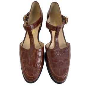 May include: A pair of brown leather t-strap heels with a crocodile texture. The shoes have a rounded toe, a buckle closure, and a small heel. The shoes are displayed on a white background.