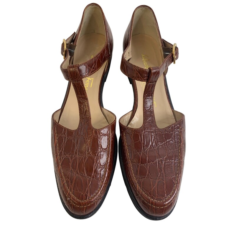 May include: A pair of brown leather t-strap heels with a crocodile texture. The shoes have a rounded toe, a buckle closure, and a small heel. The shoes are displayed on a white background.