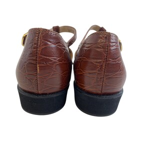May include: Pair of brown leather Mary Jane shoes with a textured, crocodile-like pattern. The shoes have a black sole and a gold-colored buckle on the strap. The shoes are viewed from the back.