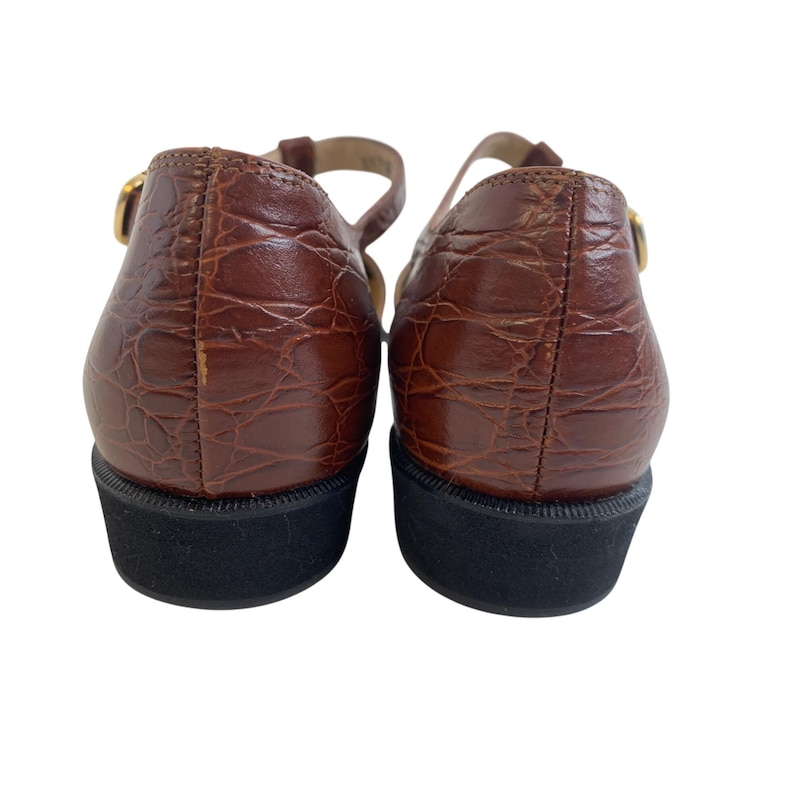 May include: Pair of brown leather Mary Jane shoes with a textured, crocodile-like pattern. The shoes have a black sole and a gold-colored buckle on the strap. The shoes are viewed from the back.