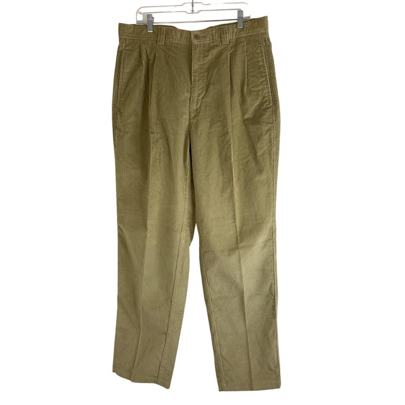 May include: Tan corduroy pants with a pleated front, hanging on a white hanger. The pants have a button and zipper closure, and pockets. The corduroy fabric has vertical ridges.