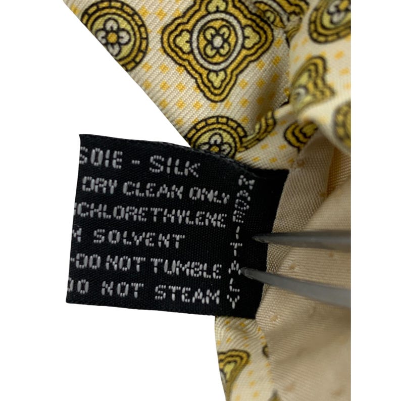 May include: Close-up of a silk tie with a repeating yellow and cream-colored floral pattern. The tag reads "SOIE - SILK" and "DRY CLEAN ONLY". The tie is held by a pair of silver tweezers.