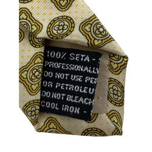 May include: Close-up of a silk tie with a repeating yellow and black floral pattern on a cream background. The label reads "100% SETA - PROFESSIONALLY DO NOT USE PER OR PETROLEU DO NOT BLEACH COOL IRON -"