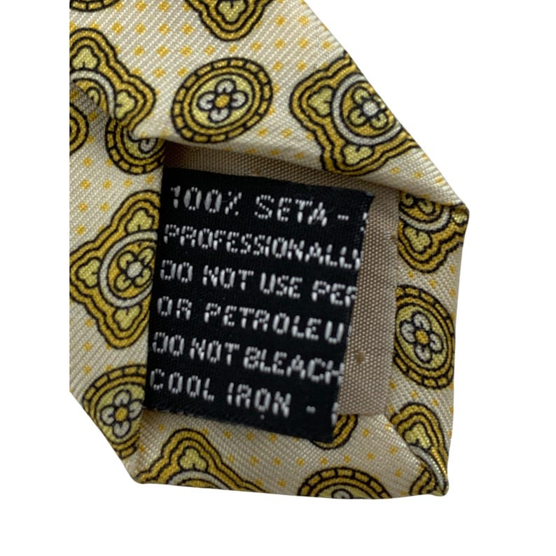 May include: Close-up of a silk tie with a repeating yellow and black floral pattern on a cream background. The label reads "100% SETA - PROFESSIONALLY DO NOT USE PER OR PETROLEU DO NOT BLEACH COOL IRON -"