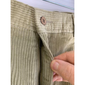 May include: Close-up of light green corduroy pants. The pants have a button closure and a zipper. The corduroy fabric has vertical ridges. A hand is pulling the zipper down.