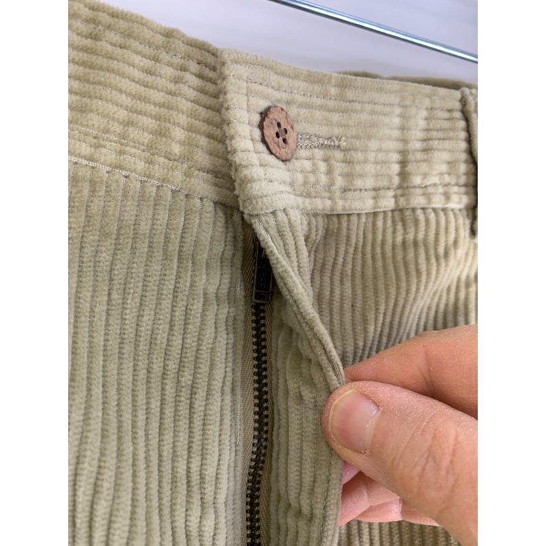 May include: Close-up of light green corduroy pants. The pants have a button closure and a zipper. The corduroy fabric has vertical ridges. A hand is pulling the zipper down.