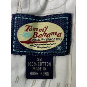May include: Close-up of a navy blue Tommy Bahama label. The label features the brand name in a stylized font, a logo with a sailboat and waves, and the text "QUALITY SINCE 1993." Below the label is a tag with the size 38, and the text "100% COTTON MADE IN HONG KONG."