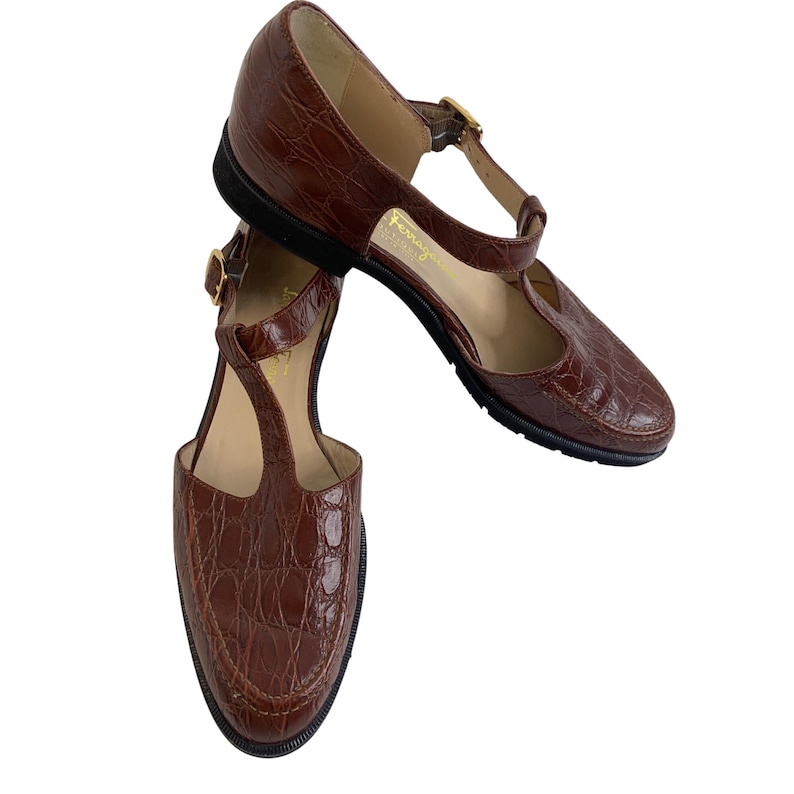 May include: Pair of brown leather T-strap shoes with a crocodile texture. The shoes have a rounded toe, a strap that goes across the top of the foot, and a buckle closure. The soles are black, and the shoes appear to be vintage.