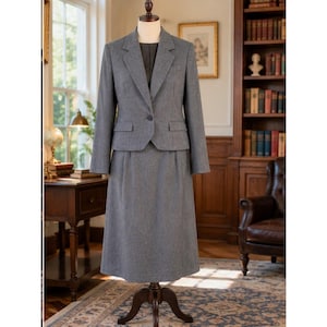 May include: A grey two-piece suit set displayed on a mannequin, consisting of a single-breasted blazer with a notched lapel, a single front button, and two flap pockets, paired with a matching mid-length skirt featuring a straight silhouette.
