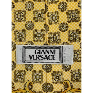 May include: Close-up of a yellow silk tie with a repeating geometric pattern in black and gold. The tie features the "GIANNI VERSACE" label in white with black text. The tie is made in Italy.