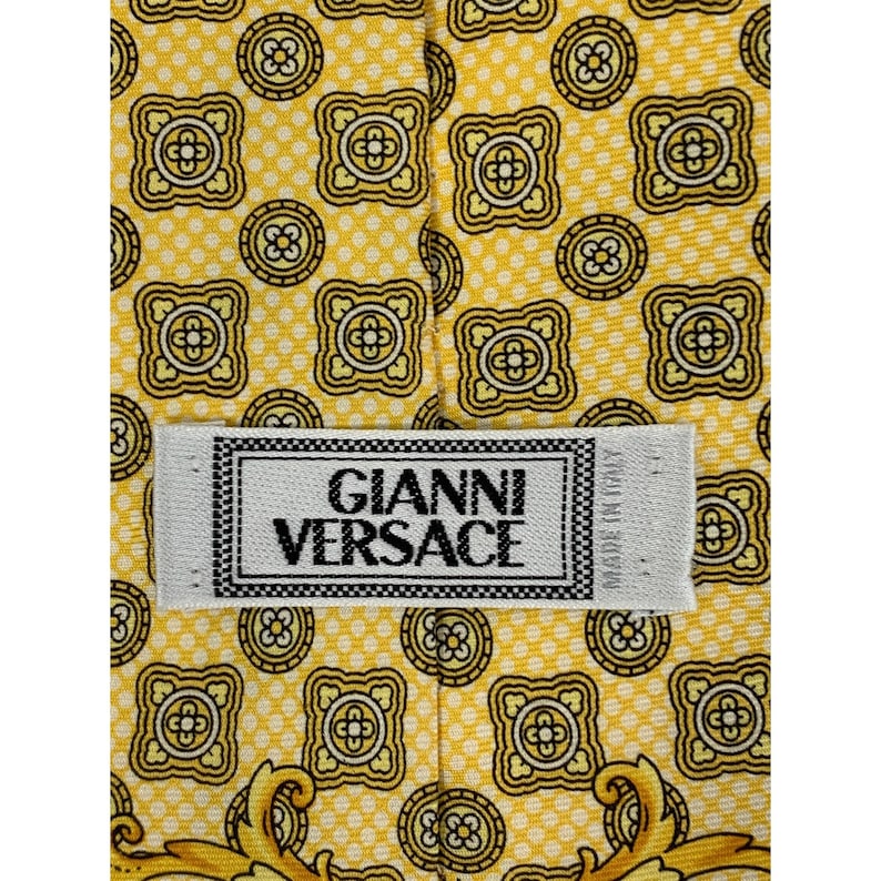 May include: Close-up of a yellow silk tie with a repeating geometric pattern in black and gold. The tie features the "GIANNI VERSACE" label in white with black text. The tie is made in Italy.