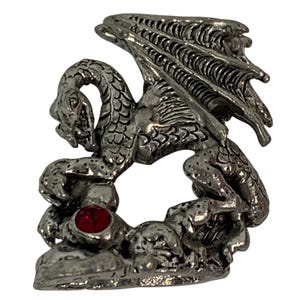 May include: A silver-colored dragon figurine with detailed scales, wings, and a red gemstone eye. The dragon is perched on a rocky base, showcasing intricate design and texture. This fantasy-themed collectible is suitable for display.