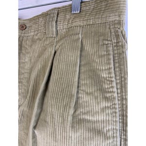 May include: Close-up of tan corduroy pants. The pants have a button closure, belt loops, and pleated front. The corduroy fabric has vertical ridges. The pants are hanging on a metal hanger.