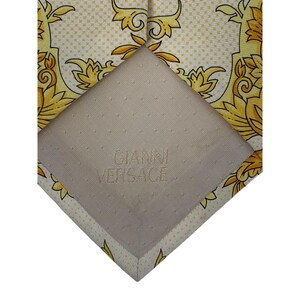May include: Close-up of a yellow and white patterned tie with a repeating floral design. The tie's underside is beige and features the text "GIANNI VERSACE" in a subtle embossed font.