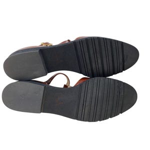 May include: Brown leather sandals with a T-strap design and a buckle closure. The sandals have a textured, dark brown sole. The image shows the bottom of the sandals, revealing the tread pattern.