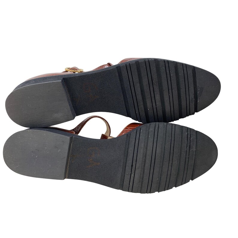 May include: Brown leather sandals with a T-strap design and a buckle closure. The sandals have a textured, dark brown sole. The image shows the bottom of the sandals, revealing the tread pattern.