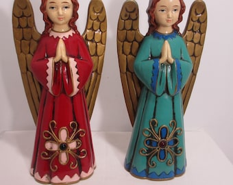 May include: Two angel figurines with folded hands. One angel wears a red robe with pink trim and a gold floral design. The other angel wears a teal robe with blue trim and a gold floral design. Both have gold wings and red hair.