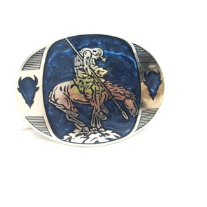 May include: Oval silver-tone belt buckle with a Native American warrior on horseback, holding a spear. The background is a swirling blue enamel. Two bison silhouettes flank the central design. The buckle is a Western-themed accessory.