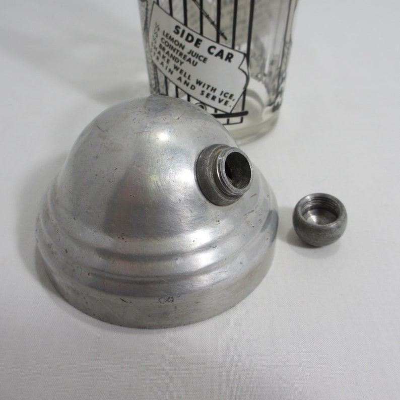 May include: A silver cocktail shaker top with a screw-on lid. The top has a small hole in the centre. The shaker top is sitting on a white surface.