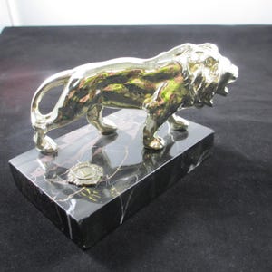 May include: A gold-colored lion statue with an open mouth, mounted on a black marble base. The base has white veining and a small, circular emblem. The lion is in a roaring pose, showcasing its teeth.