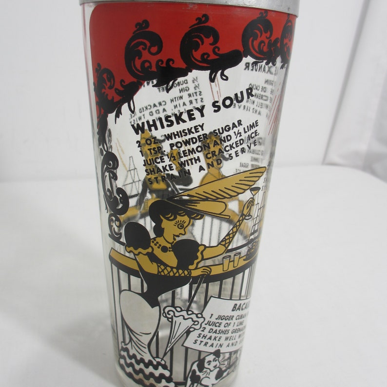 May include: A vintage glass cocktail shaker with a red, black and gold design. The shaker features a recipe for a "Whiskey Sour" with a whimsical illustration of a woman in a hat and a cat.