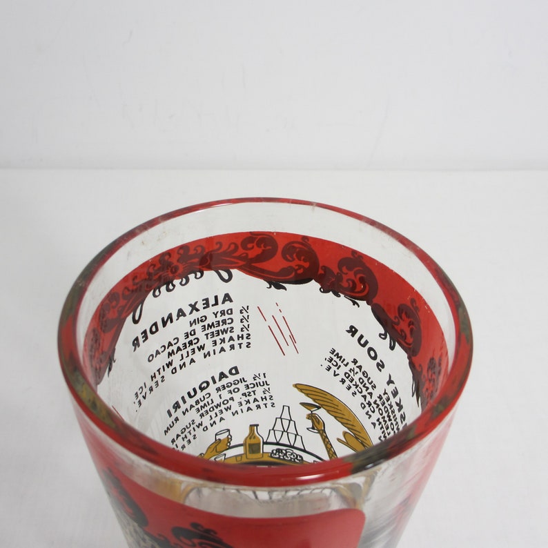 May include: A clear glass cocktail shaker with a red rim and a red and white design. The shaker has instructions for making a Daiquiri and an Alexander cocktail printed on the side.