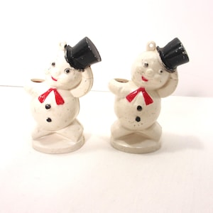 May include: Two vintage snowman figurines. Each snowman is off-white with a black top hat, red scarf, and black button details. The snowmen are holding their hats and have a smiling face.