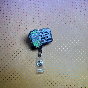 May include: A badge reel with a rectangular design featuring a gumball machine illustration. The text "IT'S OK TO HAVE MIXED EMOTIONS" is displayed on a light blue background. The badge reel has a clear plastic strap and a black cord.