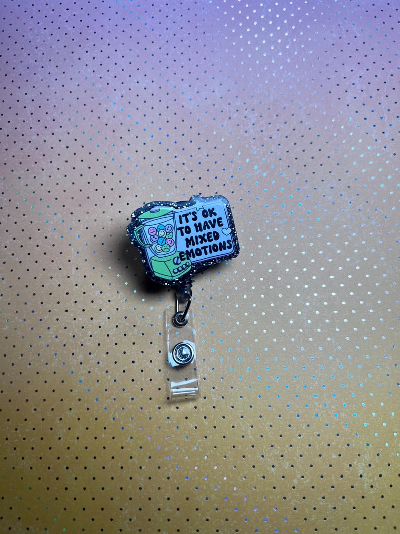 May include: A badge reel with a rectangular design featuring a gumball machine illustration. The text "IT'S OK TO HAVE MIXED EMOTIONS" is displayed on a light blue background. The badge reel has a clear plastic strap and a black cord.