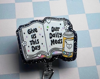 May include: A badge reel with a glittery black border. The badge features an open book with the words "Give us This Day" on the left page and "Our Daily Meds" on the right. A pill bottle with a heart is on the right side. The background is a light blue and white checkered pattern.