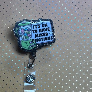 May include: A retractable badge reel with a black glitter border. The badge reel features a cartoon blender filled with smiley faces and the text "IT'S OK TO HAVE MIXED EMOTIONS". The background is a light brown color with silver polka dots.