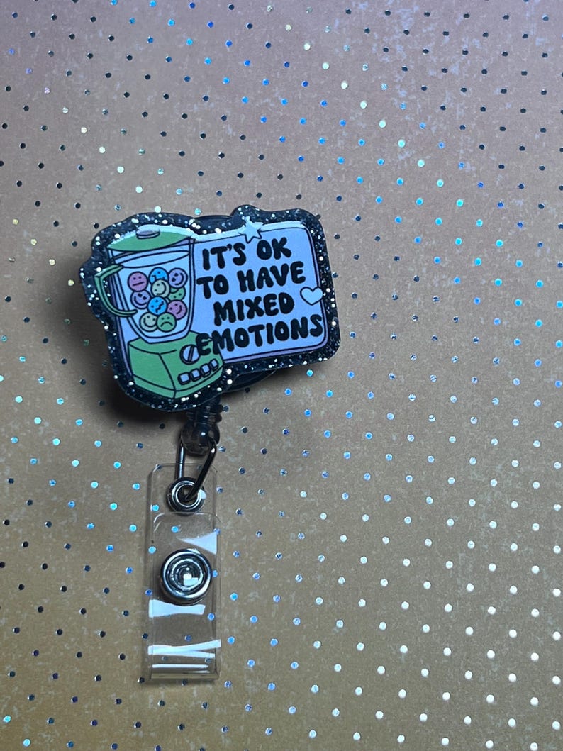 May include: A retractable badge reel with a black glitter border. The badge reel features a cartoon blender filled with smiley faces and the text "IT'S OK TO HAVE MIXED EMOTIONS". The background is a light brown color with silver polka dots.