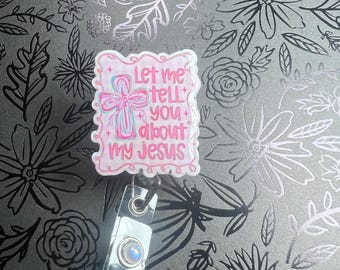May include: A pink and white badge reel with the text "Let me tell you about my Jesus." The badge has a cross design with blue and pink accents. The reel has a silver clip and a clear plastic strap.