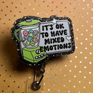 May include: A badge reel with a black glitter border. The badge features a green blender filled with smiley faces and the text "IT'S OK TO HAVE MIXED EMOTIONS". The background is orange with white dots.