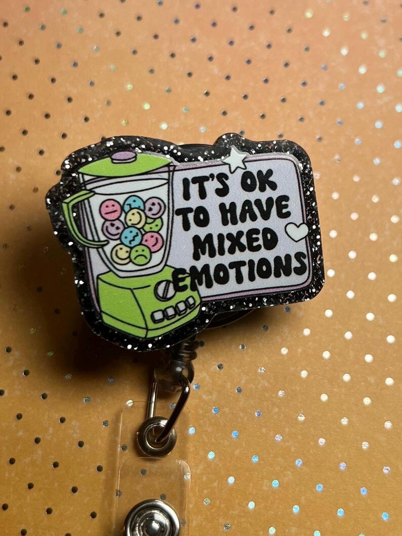 May include: A badge reel with a black glitter border. The badge features a green blender filled with smiley faces and the text "IT'S OK TO HAVE MIXED EMOTIONS". The background is orange with white dots.