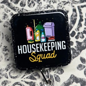 Puede incluir: Insignia negra con las palabras "HOUSEKEEPING Squad" en blanco y amarillo. La insignia presenta ilustraciones de productos de limpieza, incluyendo un pulverizador, un cubo de basura y una botella de limpiador.