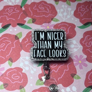 May include: A black and silver badge reel with the text "I'M NICER THAN MY FACE LOOKS" in white. The badge has a silver glitter border and a retractable cord with a clear plastic strap.