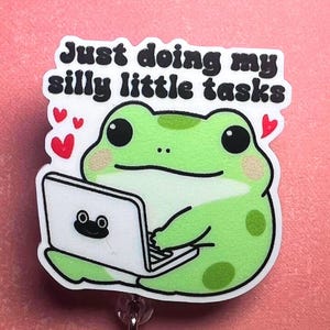 Może przedstawiać: Odznaka z zieloną żabą z kreskówki używającą laptopa, z napisem "Just doing my silly little tasks" powyżej. Żaba ma duże czarne oczy i się uśmiecha. Obecne są również czerwone serca.