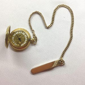 May include: A gold-toned pocket watch with an open lid, revealing a white face with black Roman numerals. The watch is attached to a gold chain and a gold-toned tie clip. The watch and chain are on a white surface.