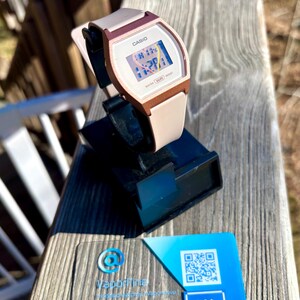 May include: A Casio digital watch with a white face and a pink and brown strap. The watch is displayed on a black stand. A business card with a blue and white design is in the foreground.