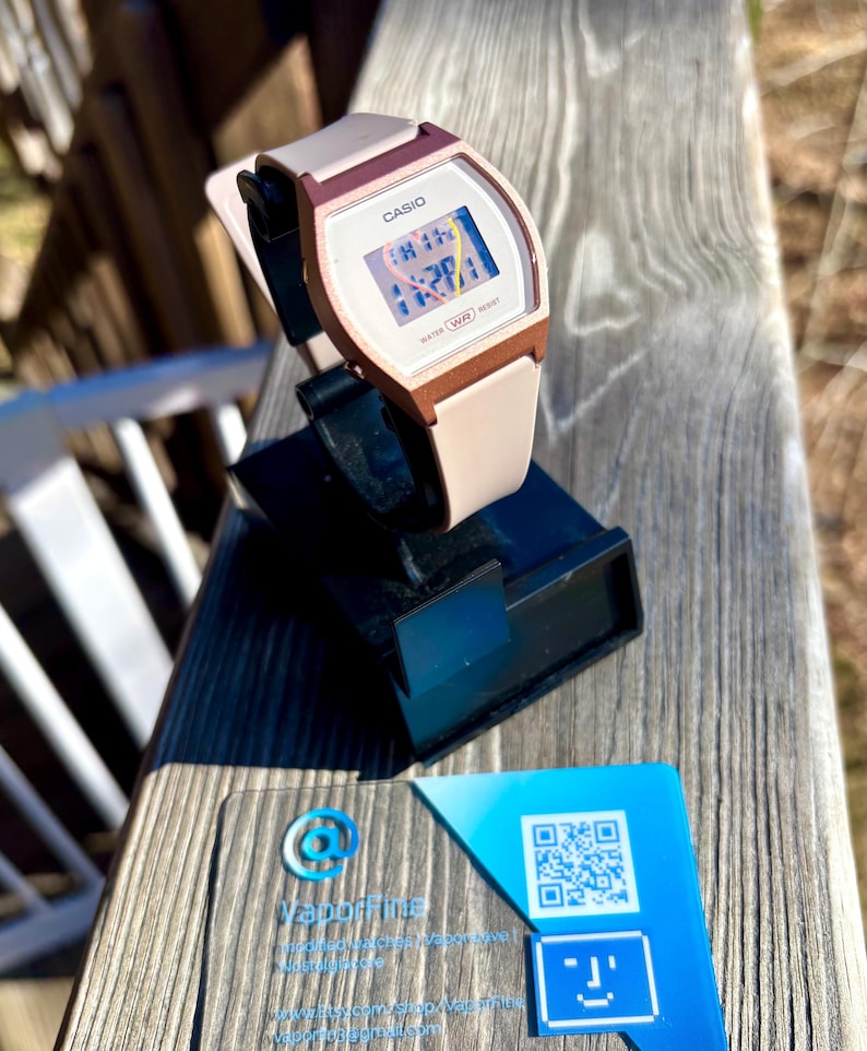 May include: A Casio digital watch with a white face and a pink and brown strap. The watch is displayed on a black stand. A business card with a blue and white design is in the foreground.
