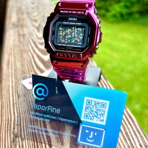 Pode incluir: Um relógio digital rosa vibrante com um visor preto mostrando pontos coloridos. O relógio apresenta a marca "SKMEI" e vários botões de função. Um cartão de visita com o logotipo "VaporFine" e informações de contato está na frente do relógio.