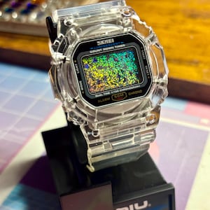 K&ouml;nnte beinhalten: Transparente digitale SKMEI-Uhr mit schwarzem Zifferblatt und farbenfroher, glitzernder Anzeige. Die Uhr hat ein transparentes Kunststoffarmband und -geh&auml;use. Funktionen: Countdown-Timer, Alarm und Chronograph. Die Uhr steht auf einem schwarzen St&auml;nder.