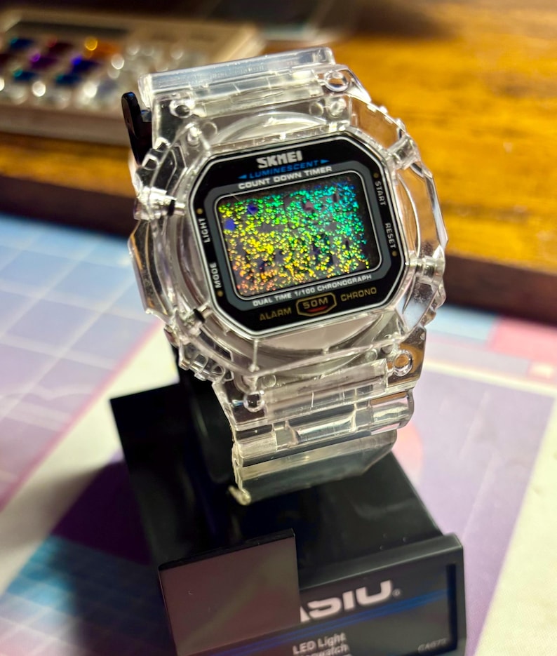 K&ouml;nnte beinhalten: Transparente digitale SKMEI-Uhr mit schwarzem Zifferblatt und farbenfroher, glitzernder Anzeige. Die Uhr hat ein transparentes Kunststoffarmband und -geh&auml;use. Funktionen: Countdown-Timer, Alarm und Chronograph. Die Uhr steht auf einem schwarzen St&auml;nder.