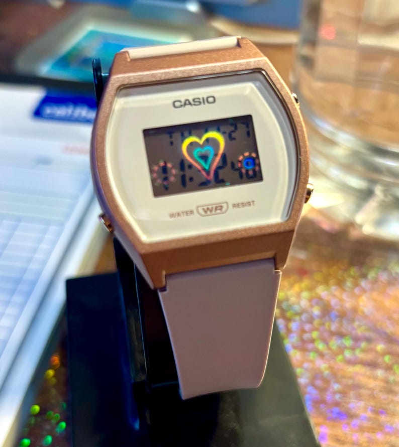 May include: A Casio digital watch with a rose gold case and a white face. The digital display shows the time and a colourful heart graphic. The watch has a light pink strap and the text "WATER WR RESIST".