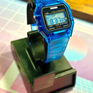 Puede incluir: Un reloj digital azul transparente con una esfera negra que muestra la hora 8:37:09. El reloj tiene una correa azul a juego y est&aacute; etiquetado como "SKMEI". Incluye "ALARM CHRONOGRAPH", "50M RESIST" y "WATER".