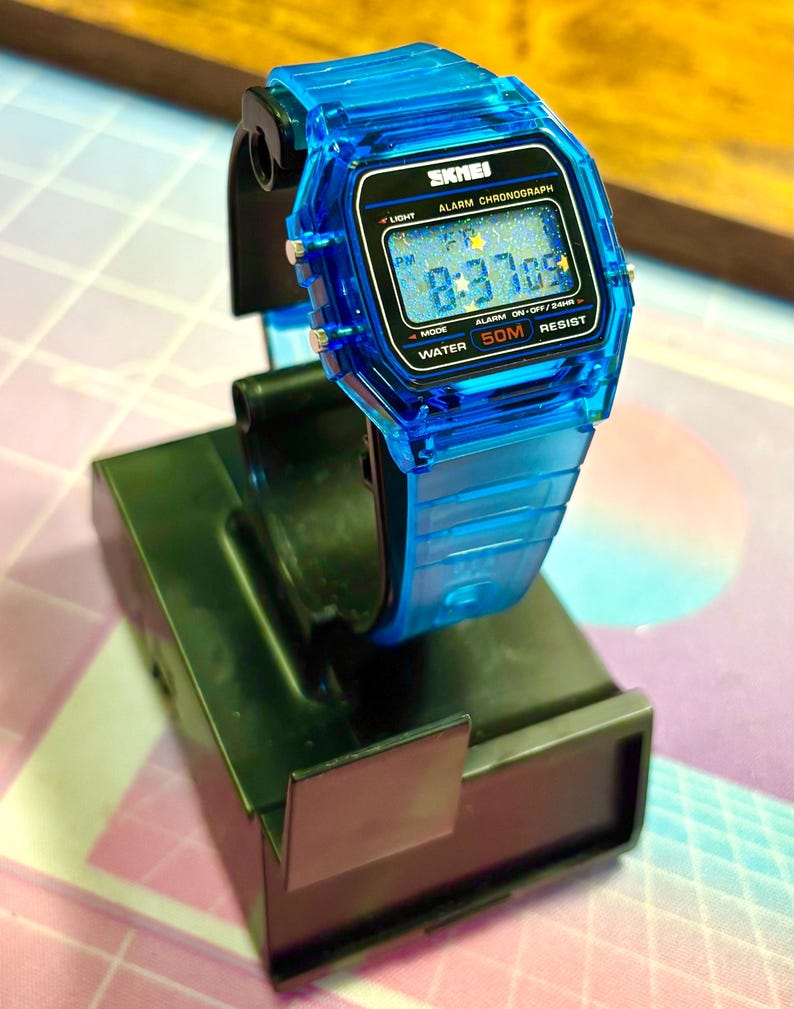 Puede incluir: Un reloj digital azul transparente con una esfera negra que muestra la hora 8:37:09. El reloj tiene una correa azul a juego y est&aacute; etiquetado como "SKMEI". Incluye "ALARM CHRONOGRAPH", "50M RESIST" y "WATER".