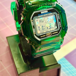 Pu&ograve; includere: Un orologio digitale SKMEI verde trasparente con un display nero che mostra l'ora e la data. L'orologio ha un quadrante quadrato ed &egrave; montato su un supporto verde scuro. Il quadrante mostra il testo "SKMEI", "LUMINESCENT", "COUNT DOWN TIMER", "DUAL TIME 1/100 CHRONOGRAPH", "ALARM 50M CHRONO".