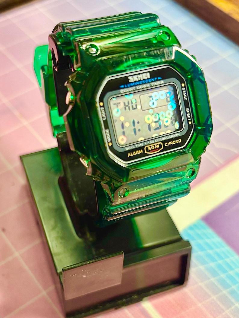 Pu&ograve; includere: Un orologio digitale SKMEI verde trasparente con un display nero che mostra l'ora e la data. L'orologio ha un quadrante quadrato ed &egrave; montato su un supporto verde scuro. Il quadrante mostra il testo "SKMEI", "LUMINESCENT", "COUNT DOWN TIMER", "DUAL TIME 1/100 CHRONOGRAPH", "ALARM 50M CHRONO".